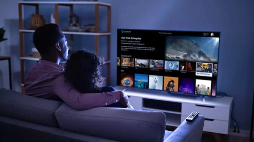 A couple sitting on a couch in a dimly lit room, watching a streaming service on their TV. The screen shows what appears to be a Star Trek: Enterprise content page with episode information and related content thumbnails. The intimate setting demonstrates how IPTV services provide at-home entertainment for couples with access to premium content like sci-fi series.