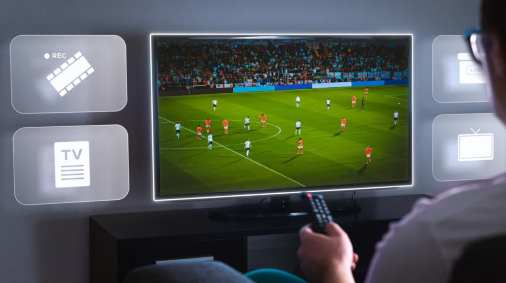 A TV screen showing a live soccer/football match with players in white and orange/red jerseys on a green field. There's a stadium crowd visible in the background. Around the screen are interface icons including "REC" (recording), "TV" (guide), and "LIVE" icons, suggesting DVR and live TV functionality. A person's hand is visible holding a remote control in the foreground.