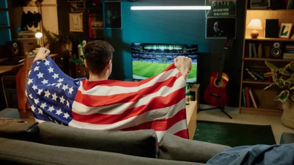 A rear view of a person sitting on a gray couch draped with an American flag while watching a soccer/football match on TV. The room appears to be a casual living space with bookshelves, a guitar, and other personal items visible. The scene conveys the idea of watching international sports content via streaming services.