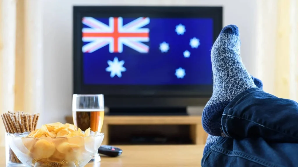 A relaxed viewing setup with someone's sock-covered feet propped up, watching a TV displaying the Australian flag. There are snacks (chips and pretzels) and a beverage visible, portraying comfortable at-home viewing of Australian content.