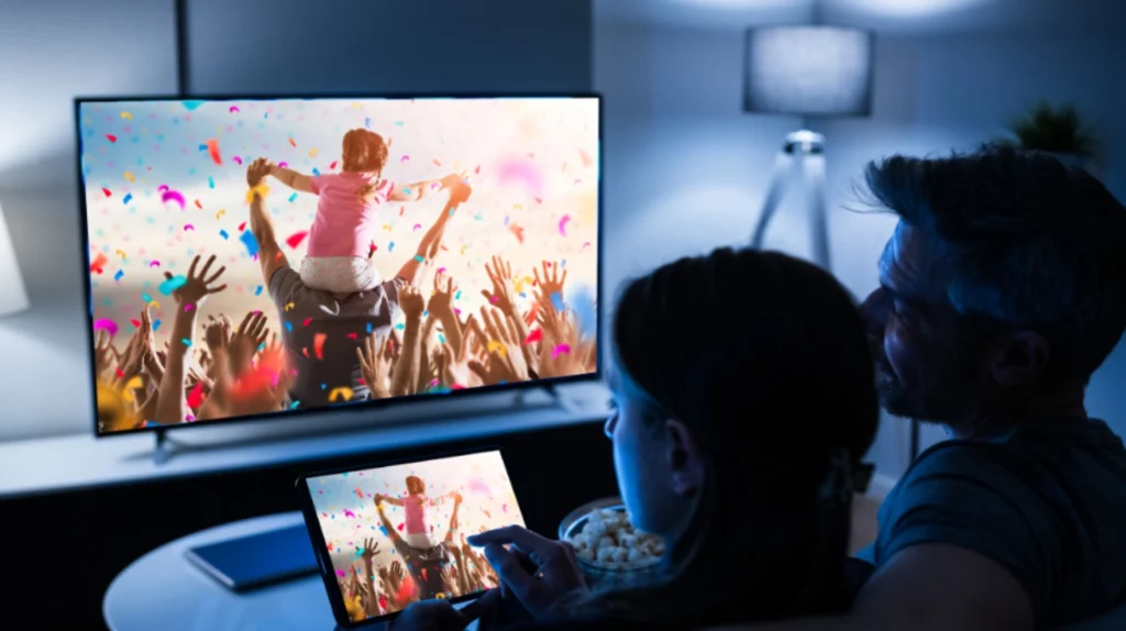 A couple watching content on both a TV and tablet simultaneously. The TV and tablet display the same vibrant scene showing a celebration with confetti and a child on someone's shoulders. They're enjoying popcorn in a dimly lit room, demonstrating multi-device streaming capabilities.