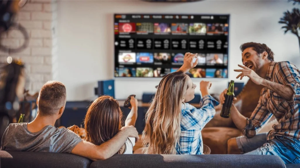 A group of friends sitting on a couch, enjoying entertainment on a large TV screen. They appear excited and engaged, with some holding beverages. The TV display shows what looks like a streaming service interface with multiple content thumbnails arranged in a grid pattern. This image illustrates the social aspect of streaming entertainment.